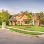 Elegant stone family home with manicured lawn, trees, and driveway on a sunny day.